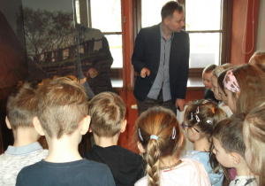 Pan Arkadiusz kustosz muzeum prezentuje dzieciom eksponaty z wystawy archeologicznej (stara studnia, kafle, cegły, przedmioty).